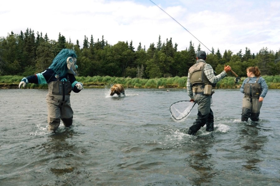 This image taken from video provided by the Seattle Kraken shows a brown bear splashing toward the NHL Kraken sea troll mascot known as Buoy, left, next to a fishing guide during the filming of a promotional video in a river outside Anchorage, Alaska, June 25, 2025. (Seattle Kraken via AP)