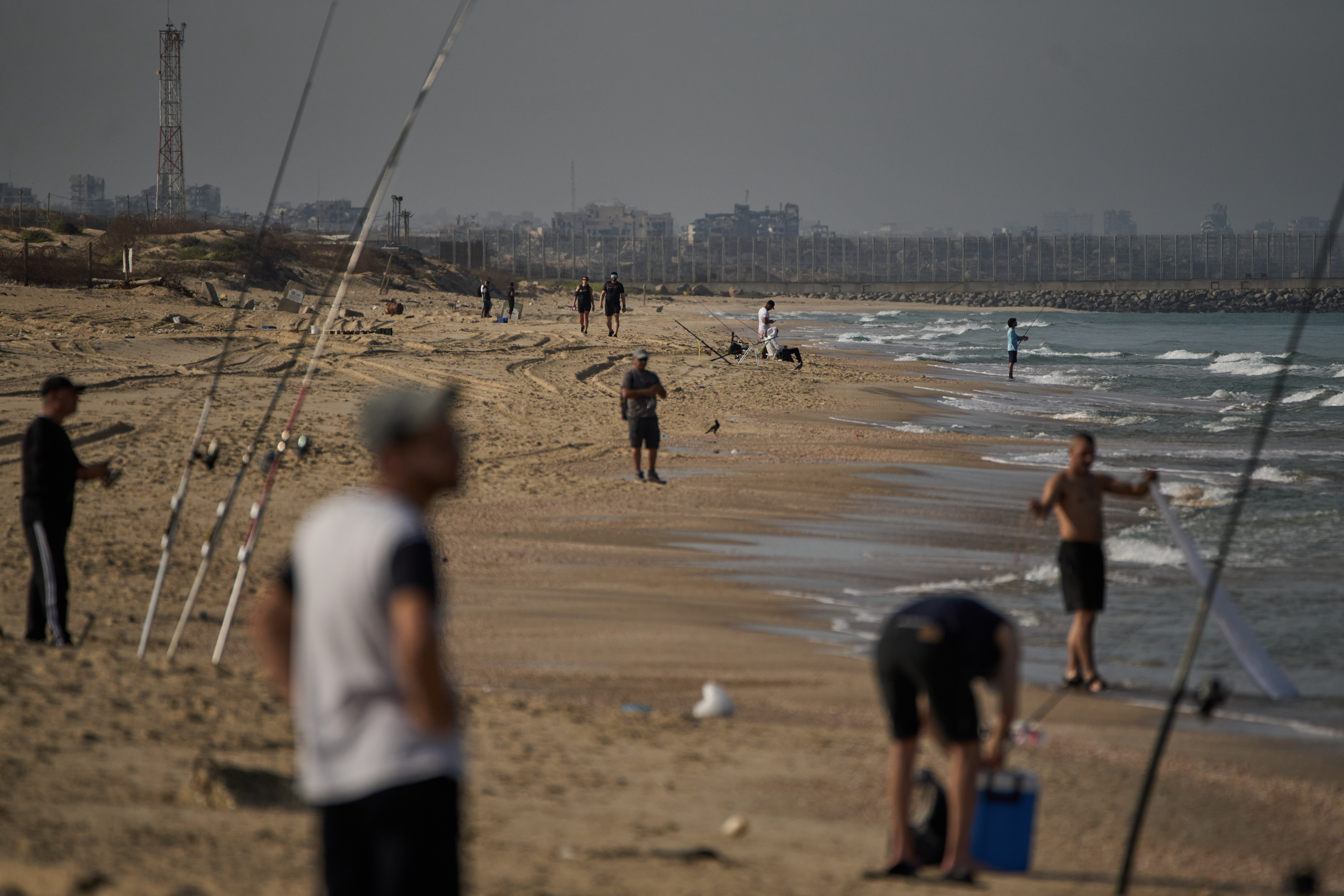 Backdropped by buildings destroyed during Israeli ground and air operations in the northern Gaza Strip, people walk and fish at Zikim Beach, near Ashkelon, in southern Israel, Friday, Oct. 17, 2025. (AP Photo/Leo Correa)