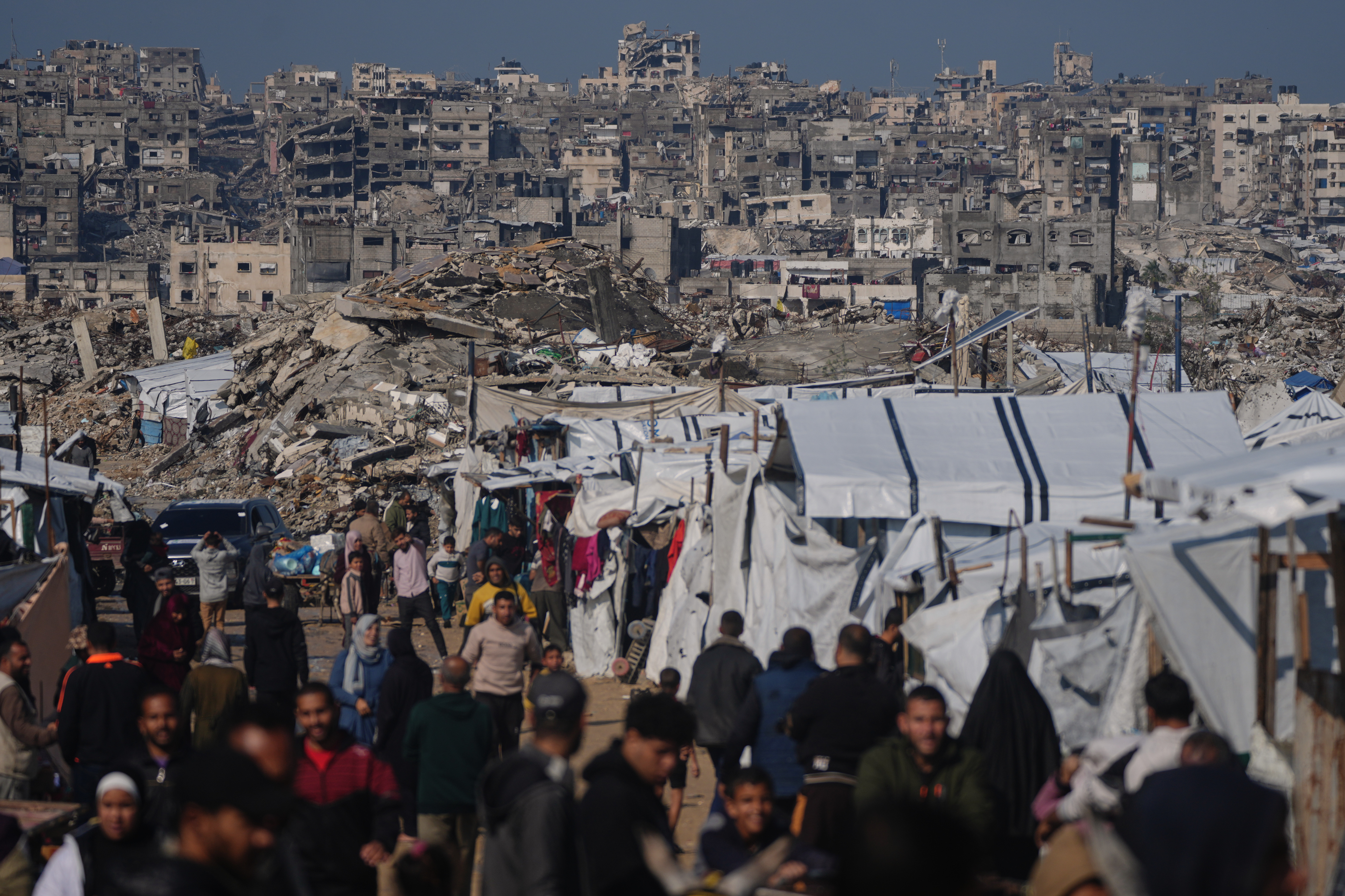 Palestinians walk along a street past a tent camp, backdropped by buildings destroyed during Israeli air and ground operations in Gaza City, Wednesday, Dec. 17, 2025. (AP Photo/Jehad Alshrafi)